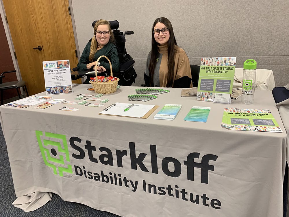 Two Starkloff team members smile behind a table filled with Access U information and promotional gifts.