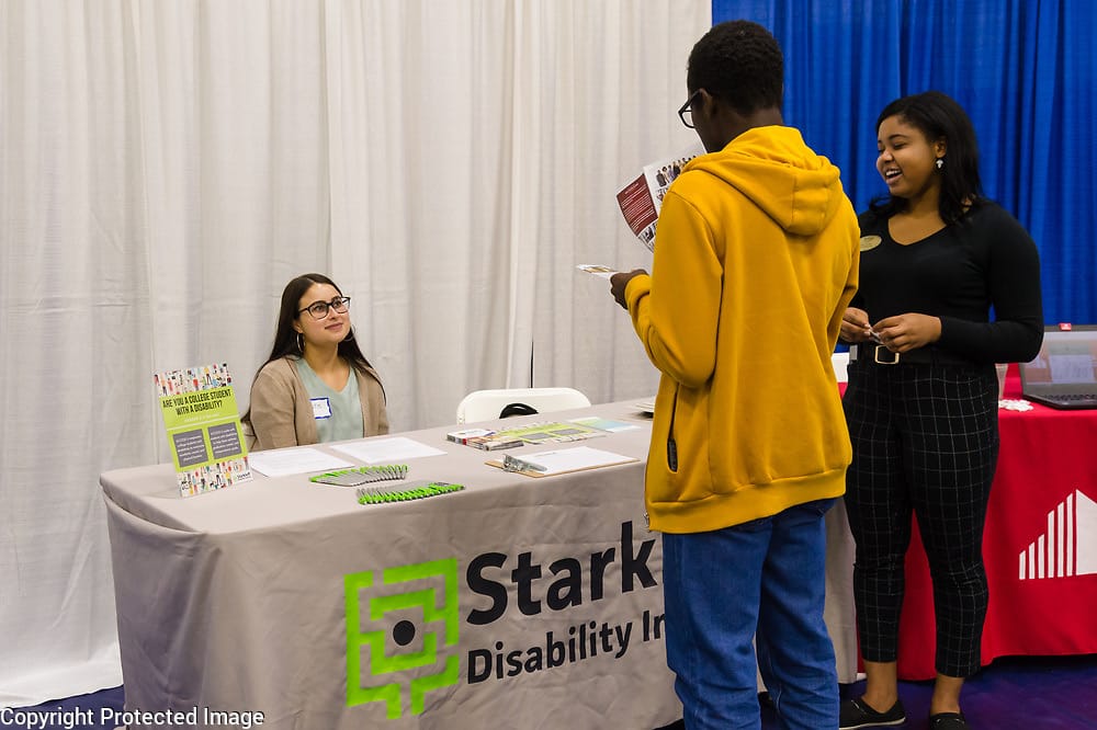 A young woman with glasses and long black hair is sitting behind a table. She is smiling at a group of people looking at Starkloff brochures.